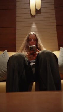 Kanepede telefonuna bakan genç bir kadının dikey videosu. Yukarıdaki lambadan gelen sıcak ve bastırılmış ışık sıcak atmosfere katkı sağlar..