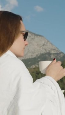 Dikey video. Açık bir sabahta balkonda uzun saçlı bir esmer kahve içer ve dağları ve ormanı sever..