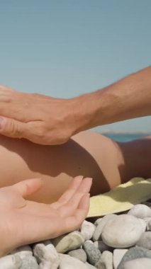 Dikey video. Bir adamın eli, kumsalda uzanmış genç bir kadının yumuşak kalçalarına güneş kremi sürer..