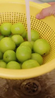 Dikey video. Mutfaktaki musluktan akan bir su deresinin altında meyveleri yıkamak için elek içinde yeşil kiraz erikleri..