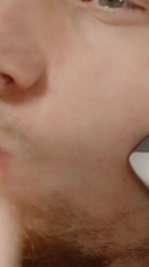 Dikey video. Yanak Arkası ve Önü Elektrikli Ustura Kullanan Genç Adam, Yavaş Hareket. Banyodaki Ayna 'da Sabah Rutini.