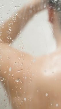 Dikey video. Sabunlu su damlası kaplı camın arkasındaki duşta çıplak bir kadının bulanık silueti, Vücudundan süzülen kamera.