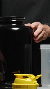 Dikey video. Bir adam, büyük bir kavanozdan bir kaşık protein tozu alır ve onu bir sallayıcının içine döker. Çeşitli Spor Hazırlıkları Yakınlarda Uzanıyor.