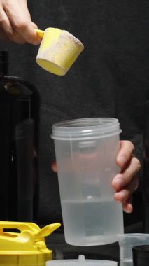 Dikey video. Çikolatalı Protein tozu bir Shaker 'a suyla doldurulur. Masanın üzerinde sayısız takviye madde olan siyah kutular.