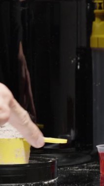 Dikey video. Masadaki çeşitlilikten hangi spor ilavesini alacağına karar veremeyen bir adam ölçü kabında kırmızı bir içecek seçer..