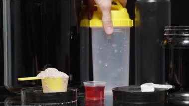 Masanın üzerinde spor malzemeleri olan siyah teneke kutular, bir adam bir kaşık protein tozu ve onu hazırlamak için bir titretici alır..