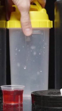 Dikey video. Masanın üzerinde spor malzemeleri olan siyah teneke kutular, bir adam bir kaşık protein tozu ve onu hazırlamak için bir titretici alır..