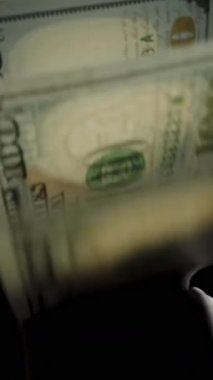 Dikey video. Elinde bir yığın para, yüz dolarlık banknotlar rüzgarda dalgalanan siyah bir arka planda, yakın planda.