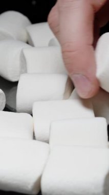 Dikey video. Beyaz Marshmallow 'u İnceleyen Erkek El, Siyah Bir Arkaplan Karşısında Masaya Bir Çok Şekerleme Dağıldı.