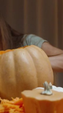 Dikey video. Uzun saçlı bir esmer balkabağına gözlerini oyuyor. Sıcak bir akşamda Tatil Öncesi Sıcak Ev Ayarları.