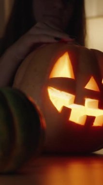 Dikey video. Karanlık bir odada, bir kadın Jack-O 'Lantern' in Lid 'ini açar ve içeri bakar, Yüzü mum ışığında ışıl ışıl parlar.