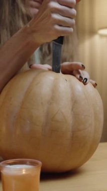 Dikey video. Genç Uzun Saçlı Kadın, Balkabağından Bıçağı Çekmek İçin Mücadele Ediyor, Bir Jack-o 'Lantern Yaratıyor. Önce Konforlu Ev Ayarları