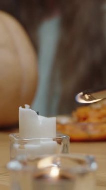 Dikey video. Cadılar Bayramı 'nda Balkabakları Arasındaki Masada Bir Mum yakmak. Arka planda, bıçaklı bir fener oymaya hazır bir kadın var. Yavaş Hareket.