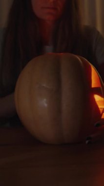 Dikey video. Karanlık Oda 'da, Kadın Masada Otururken Balkabağı Jack-o' -Fener Suratını Kameraya Çeviriyor. İçerideki Parlak Alevli Işık, Lid 'le kaplıyor.