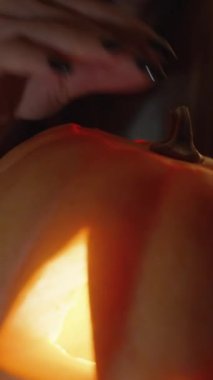 Dikey video. Balkabağı fenerinin içine bakan genç kadın, parlak mum ışığıyla aydınlatılıyor. Cadılar Bayramı Gecesi.