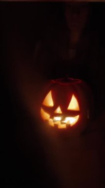 Dikey video. Karanlık bir odada, uzun saçlı bir kadın elinde bir balkabağı tutuyor aynanın önünde, arkadan görünüşü, tüyler ürpertici atmosferi, balkabağından parlak bir ışık.