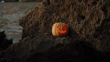 Deniz kenarında gün batımı ve Cadılar Bayramı Kutlaması. Balkabağı Jack-O-Lantern Su kenarında bir kayanın üzerinde duruyor ve parlıyor..