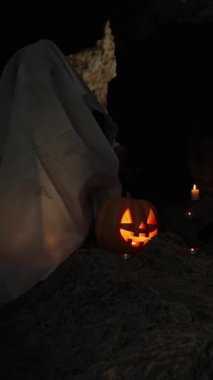 Dikey video. Beyaz çarşaflı bir hayalet cadılar bayramı gecesinde Karanlık Mağarada oturur kayalarda balkabağı, duvarlarda mumlar.