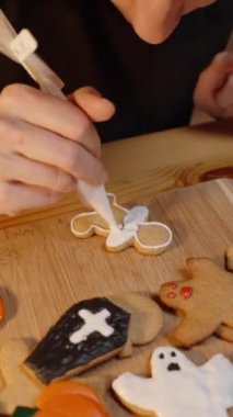 Dikey video. Yakınlaştır, bir kadın erkek şekilli kurabiyelerden birine ayrıntı eklemek için beyaz krema kullanıyor. Kurabiyelerin Cadılar Bayramı 'yla ilgili çeşitli şekilleri ve dekorasyonları var, buna balkabakları, hayaletler ve haçlar da dahil..