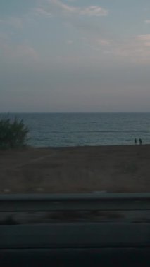 Dikey video. Hareket eden bir arabanın penceresinden manzara görüntüsü. Ön planda bariyerli bir yol var. Arka planda ise Günbatımının yumuşak ışığıyla aydınlatılan kıyı ve deniz var..