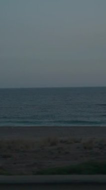 Dikey video. Önünde yol ve sebze olan hareket halindeki bir arabanın manzarası. Plaj ve okyanus ufka doğru uzanıyor. Hafif Bulutlu Gökyüzü Sahneye Huzurlu Bir Ambiance ekler.