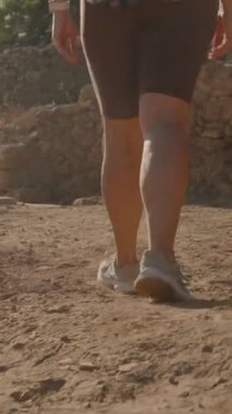 Dikey video. Batan Güneş 'in silueti altında, Antik Yol boyunca yürüyen bir kadın, Harabelerin Huzurlu Güzelliği' ne gömülmüş..