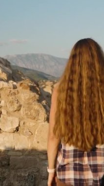Dikey video. Tarihi harabelerde ayakta duran bir kadın manzaranın güzelliğine daldırılmış Geniş Dağ Sırasına bakıyor..