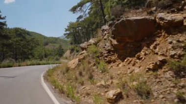 Rocky Cliffs ve Pine Trees 'in hareket halindeki bir arabadan göründüğü bir viraj yolu. İlerideki Yol Kıvrımları, Ötesinde yatan yalanlar hakkında merak kıvılcımları. Temiz Gök ve Yeşil Tepeler Bir Manzaralı ve Maceralı Atmosfer Oluşturur.