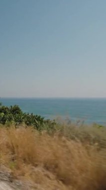 Dikey video. Ocean Views, Lush Vegetation ve Power Line 'lı bir sahil yolu. Açık Mavi Gökyüzü ve Köpüklü Su Sahilsel Sürücünün Güzelliğini Işıklandıran Manzaralı ve Huzurlu Bir Atmosfer Oluşturur.