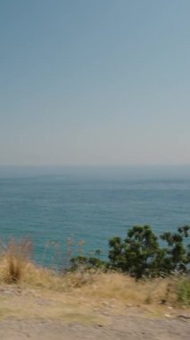 Dikey video. Bir Arabadan Sahil Manzarası Geniş Mavi Okyanusu ve Açık Gökyüzünü Gösteriyor. Yol kenarı bitkisi Manzaraya Eklenir, Huzurlu ve ferahlatıcı bir atmosfer yaratır Yolculuk boyunca.