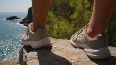 Uçurumun kenarında dikilen, spor ayakkabı giyen bir adam. Uzakta Okyanus ve Kayalar, Doğal Manzarayı Keşfetmenin Macerası ve Heyecanını Vurguluyor, Çarpıcı bir görünüm oluşturur..