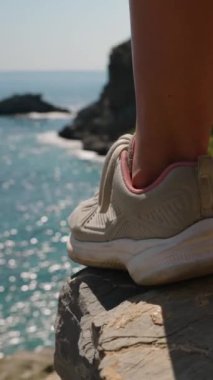 Dikey video. Uçurumun kenarında dikilen, spor ayakkabı giyen bir adam. Uzakta Okyanus ve Kayalar, Doğal Manzarayı Keşfetmenin Macerası ve Heyecanını Vurguluyor, Çarpıcı bir görünüm oluşturur..