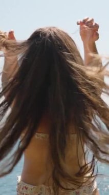 Dikey video. Bir kadın, bir uçurumun kenarında dikilirken uzun saçlarını ağır çekimde savuruyor. Okyanus ve berrak mavi gökyüzü çarpıcı bir zemin sağlar, bir sahne ortamındaki özgürlük ve doğal güzelliği yakalar..