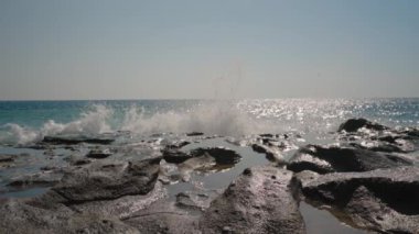 Yavaş çekimde Rocky Shoreline 'a düşen dalgalar. Güneş ışığı sudan yansıyarak ışıltılı efektler yaratır. Açık Mavi Gökyüzü Huzurlu ve Görkemli Kıyı Sahnesini Geliştirir.