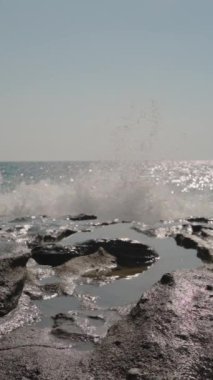 Dikey video. Yavaş çekimde Rocky Shoreline 'a düşen dalgalar. Güneş ışığı sudan yansıyarak ışıltılı efektler yaratır. Açık Mavi Gökyüzü Huzurlu ve Görkemli Kıyı Sahnesini Geliştirir.