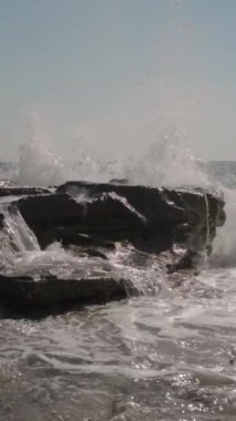 Dikey video. Dalgalar sahile çarparken Rocky Ledges 'in üzerinde çağlayan sular. Köpük ve Sis, Güneş Işığı Okyanusu ve Açık Mavi Gökyüzü tarafından geliştirilmiş dramatik, dinamik bir sahne yaratır..