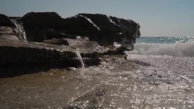 Engebeli Kayaların Üzerinde Su Şelalesi 'ne yakın çekim. Güneş ışığı nemli yüzeylere yansıyor, dokuları vurguluyor ve sakin, ışıl ışıl bir sahil atmosferi yaratıyor..