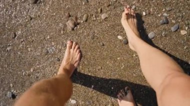 Bir adam ve bir kadın bir çakıl taşı plajında yalınayak yürüyor, Nazik Dalgalardan ayaklarını ıslatıyorlar. Yavaş Hareket Etkisi Deniz kenarındaki Dinlendirici ve Huzurlu Anı Yakalıyor.