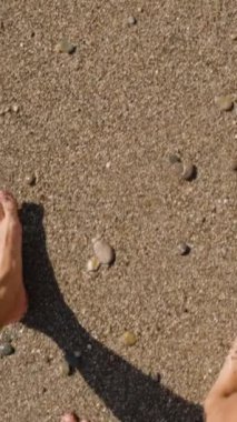 Dikey video. Çakıl Kumsalında Çıplak Ayaklı Bir Aile. Güneş ışığı, Kum yüzeyindeki adımlarını vurguluyor, Huzurlu ve rahatlatıcı bir sahne yaratıyor.