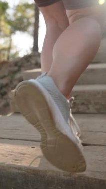 Dikey video. Adım adım, kadın ağır çekimde ahşap merdivenlere tırmanır, ağaçların arasından süzülen yumuşak güneş ışığıyla yıkanır..