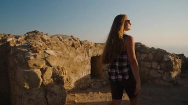 Gün batımında, bir kadın antik bir şehrin kalıntıları arasında yürüyor, altında huzur dolu bir deniz uzanıyor..
