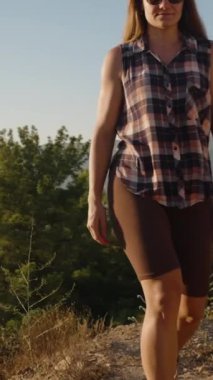 Dikey video. Bir tepe yolu boyunca emin adımlarla yürüyen kadın, Görkemli Dağ Sahnesinde Altın Gün Işığıyla yıkanır..