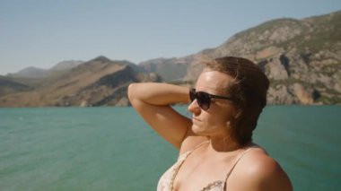 Güneş Gözlüklü Kadın Göl kenarındaki güneşli günün tadını çıkarıyor, Görkemli Dağlar ve Açık Mavi Su ile çevrili, Güneş 'ten El Kalkanı.