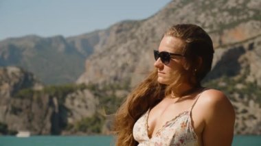 Uzun saçlı ve güneş gözlüklü düşünceli kadın uzak dağlara, dingin göle ve engebeli manzaraya bakıyor. Huzurlu bir atmosfer yaratıyor..
