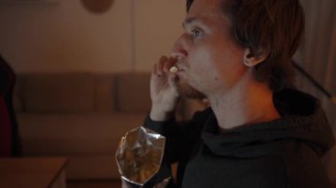 Gece atıştırması, genç bir adam cips yiyor. Samimi ışıklandırma onun rahat duruşunu vurguluyor..