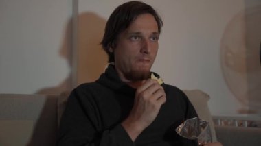 Gece atıştırması, kanepeye oturup cipslerin tadını çıkaran bir adam. Yumuşak ışık ve rahat atmosfer rahatlatıcı bir atmosfer yaratır..