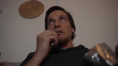 Yukarı doğru ısırıyor, ağzına bir çip sokuyor. Sahne, akşamının rahat ve rahat atmosferini yakalıyor..