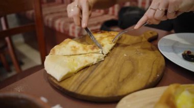 Bir dilimi keserken, genç bir kadın ustalıkla ekmeği bölüyor. Sıcak, peynirli khachapuri lezzetli bir tat vaat ediyor..