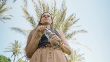 Serin içecek, genç bir kadın palmiye ağaçlarının altında su içer. Mavi gökyüzü ve yeşillik rahatlatıcı bir atmosfer yaratır..