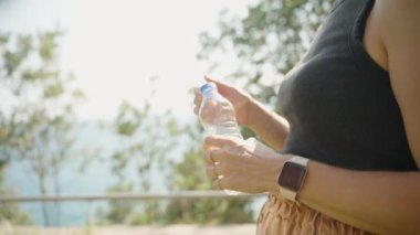 Güneşli bir günde, genç bir kadın açık havada su içer. Parlak güneş ışığı ferahlatıcı deneyime sıcaklık katar..